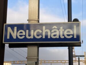 Blue and white sign reading "Neuchâtel" on a metal pole, set against a partly cloudy sky and railway tracks.