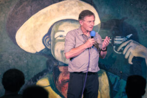 Colin Chandler speaks into a blue microphone, standing before a mural of a person with a hat and a gun. 