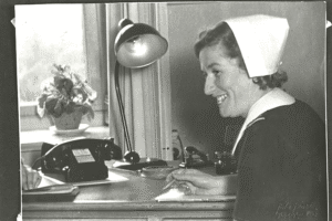 A smiling nurse sits at a desk, holding a pen. A vintage phone, desk lamp, and potted plant are in the background. The scene conveys warmth and professionalism.