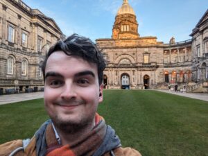 Oisin taking a selfie in front of the historic Law School building at old College, a stone building with a domed roof. The scene is set against a clear blue sky, conveying a cheerful tone.
