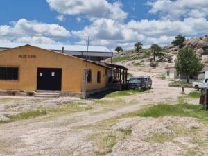 A rustic yellow building labeled "Salon Ejidal" stands under a bright blue sky with fluffy clouds. Trucks are parked nearby, with rocky terrain and trees in the background.