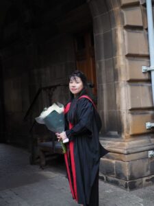 Mengying Zhang stands outside a building on graduation dressed in robes and holding a bouquet of flowerrs 