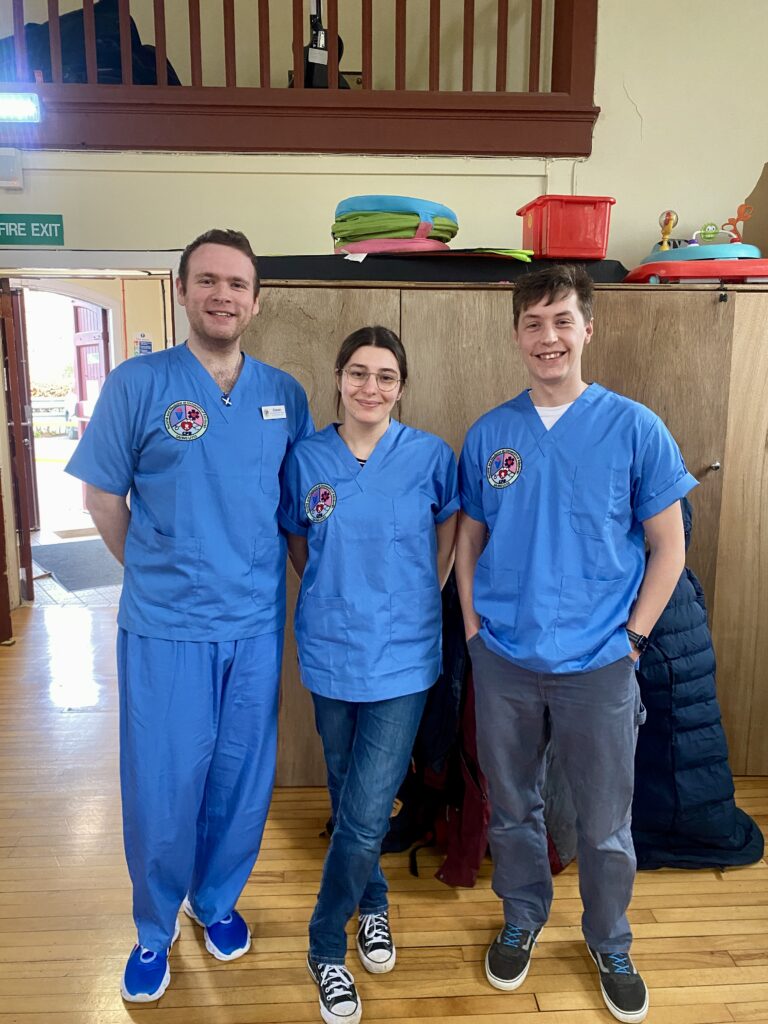 Three students stand together wearing their blue scrubs embroidered with the university logo.