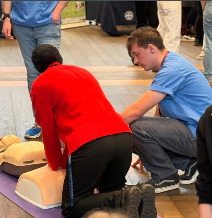A man in blue scrubs instructs a woman in a red top how to do CPR.