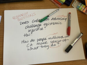 A large sheet of white paper with the following words written on it. "Research questions. Does collectrive Advocacy in Scotland challenge epistemic injustice? How? How do people (who?0 involved in CA make sense of what they do?" There are three pens on the paper, green, red black. 
