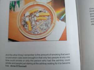 screenshot of a book: photo of a full ashtray and text saying "And the other thing I remember is the amount of smoking that went on and when rules were brought in that only two people at any one time could smoke or only the person who had the ashtray could smoke and people just staring at the ashtray waiting for it to become free." 