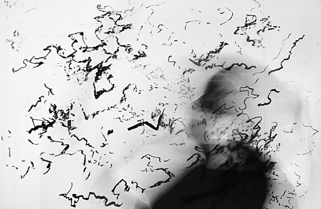 This is a contextual image for the OER. It shows in black and white a scattering of black rough short lines and dots randomly across the background. In front to the centre-right of the image is a blurred silhouette of a person. this is in dark grey and black.