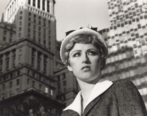 This black-and-white photograph depicts a woman with short, curled hair wearing a light-colored hat with a bow and a structured jacket with a white collar. She gazes off-camera with a confident yet contemplative expression. The background features towering buildings with geometric patterns, evoking an urban setting. The contrast between her poised demeanor and the towering architecture creates a sense of individuality and self-assuredness amidst a busy cityscape.