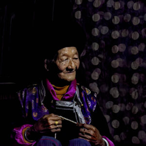 An elderly woman wearing traditional attire sits in a dimly lit setting, highlighting the intricate details of her face and hands. Her clothing features purple and gold embroidered patterns, exuding elegance. She holds a string of prayer beads in her hands, her expression calm and serene, evoking a sense of peace and introspection. The photograph emphasizes her wrinkles and the texture of her skin, conveying a feeling of timelessness and cultural depth.