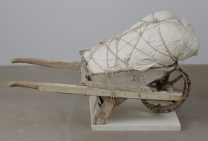 This is an art installation featuring an old wooden wheelbarrow and a large object wrapped in white fabric. The surface of the wheelbarrow shows signs of wear and tear, with the wood appearing aged and weathered, while the metal wheel is rusted and worn. The wrapped object is bulky and tightly bound with coarse rope, evoking a sense of weight and restraint. The entire piece conveys an atmosphere of labor, endurance, and the passage of time.