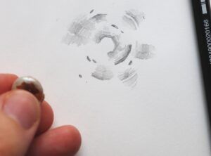 A hand holds the shell slightly above the page, where an abstract collection of lines and gradients form a black and white drawing, made with pencil, that is vaguely circular in appearance.