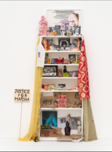 This artwork is a mixed-media installation resembling a shrine or memorial. It features a white bookshelf adorned with an assortment of photographs, small figurines, fabric, handwritten notes, and various objects.