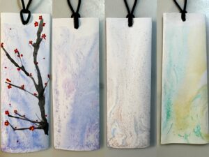 There are four bookmark works. The leftmost bookmark has a plum blossom painted on it. The background is coloured with a combination of purple and blue. The remaining three are unpainted, simply coloured bookmarks. The second from the left is coloured by a combination of blue colours, violet and gold. The third one is coloured by a combination of blue, pink and gold. The rightmost bookmark is coloured by a combination of dark green, green and gold.