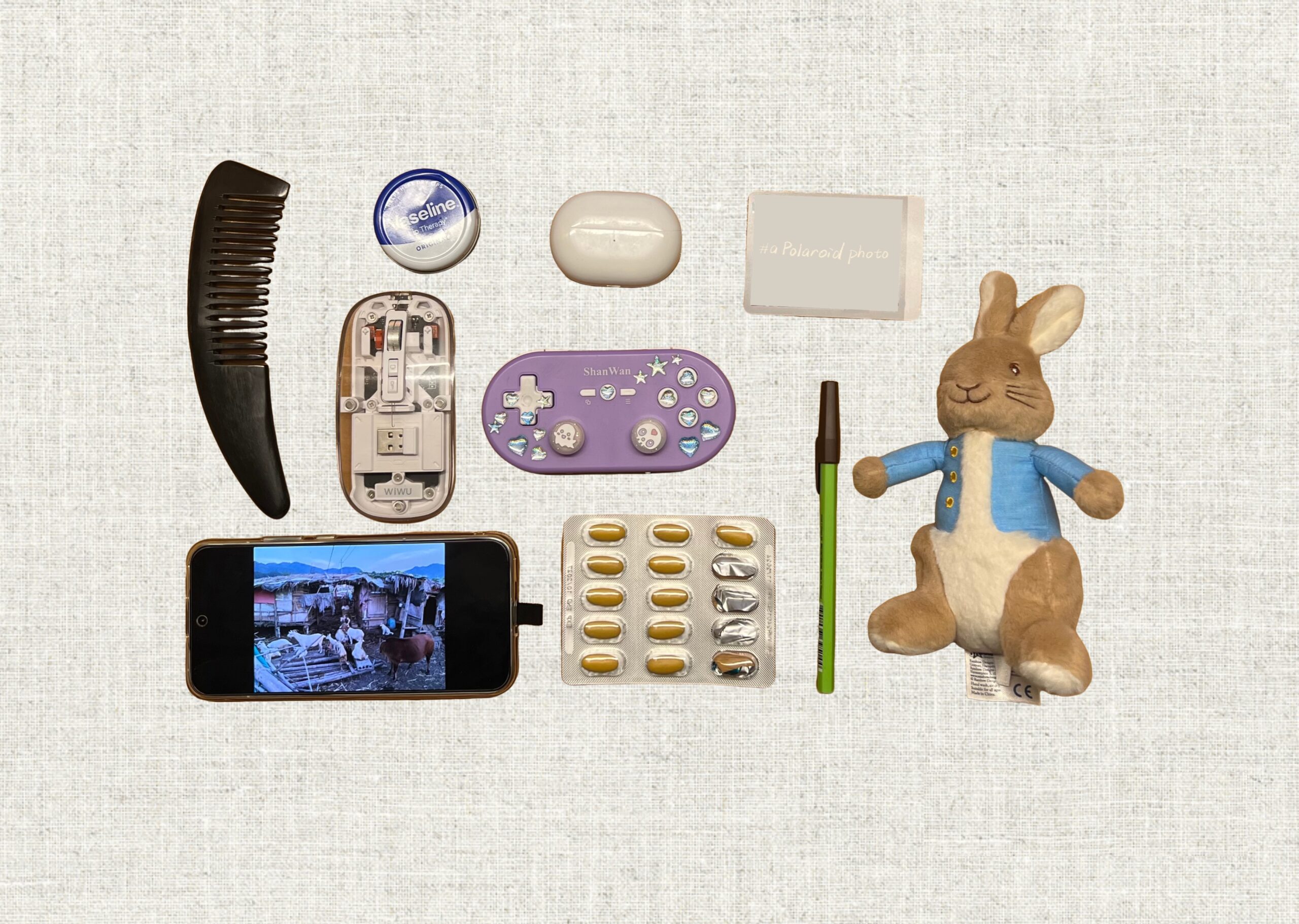 A flat lay of everyday items including a comb, game controller, phone, pen, pills, and a plush rabbit.