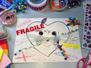This is a photo of a torn painting that has been repaired. The mended work is placed on the table surrounded by some small colored wool balls, tape rolls, ribbons, scissors, and other handmade materials needed. A crack in the top left of the heart is mended with red tape printed with the word “FRAGILE,” similar to labels used for fragile packages. The middle of the heart is sewn together with colored thread. Star stickers, heart stickers, and small colored wool balls are also used to join fragments, clearly showing the mend traces. The teardrops are filled with strings of pearls and small glitter, suggesting that these tears are precious. There are many other warm details: a white ribbon forms a bow decoration; small colored wool balls and colored paper cover the messy scribble in the top right, creating a firework-like effect; a pink paper strip covers the original “It could be better.” text, now writing “At this moment it is enough!”; a yellow paper strip covers the original “It’s so hard to get started.” on the right, now reading “Done is better than perfect.”; originally unsettling sentences are crossed out and replaced with empowering words like “My worth is defined by me!”; nearby, a handwritten sentence says “Allow yourself to take a deep breath.” The entire work uses various visible mend methods and offers diverse textures to the touch. It conveys a transformation from brokenness to gentle mending, filled with self-care. It’s like an anxious heart being carefully wrapped, comforted, and becoming warm and strong.