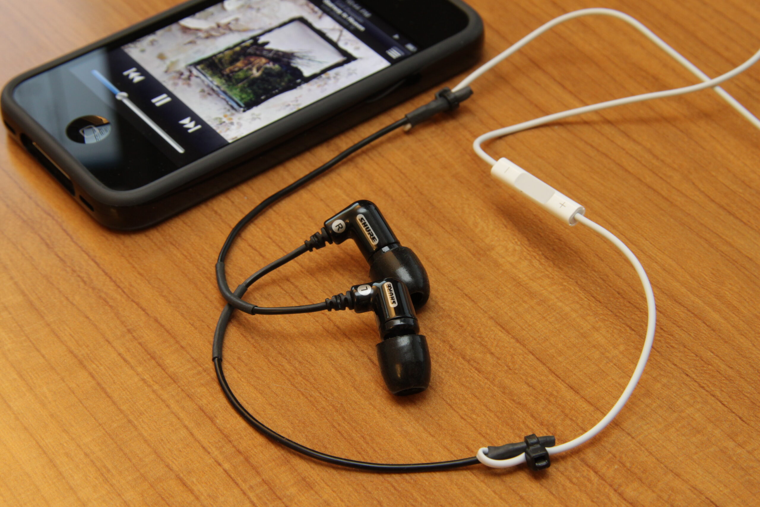 Listening Tools A mobilephone playing audio on a wooden table, with a pair of in-ear earphones placed beside it.