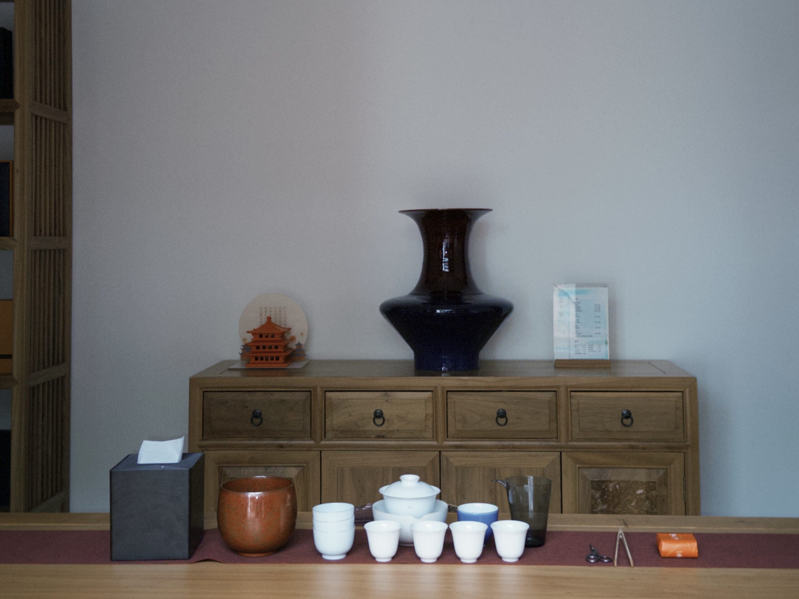 Tea Set Arrangement in a Minimal Interior A neatly arranged tea set on a wooden table, including a gaiwan, white teacups, a glass pitcher, and tea tools, placed in front of a wooden cabinet with a large blue vase, a small architectural model, and a book on display.