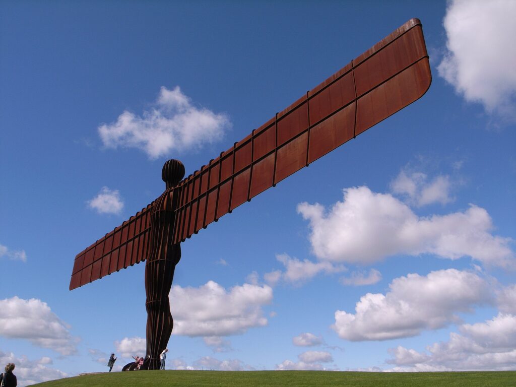 A large steel sculpture of a human figure with wide wings, standing on a green hill under a bright blue sky with clouds. People are seen walking nearby.