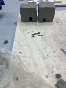 two ceramic house-like structures with flat rooves sit side by side on a wooden board.