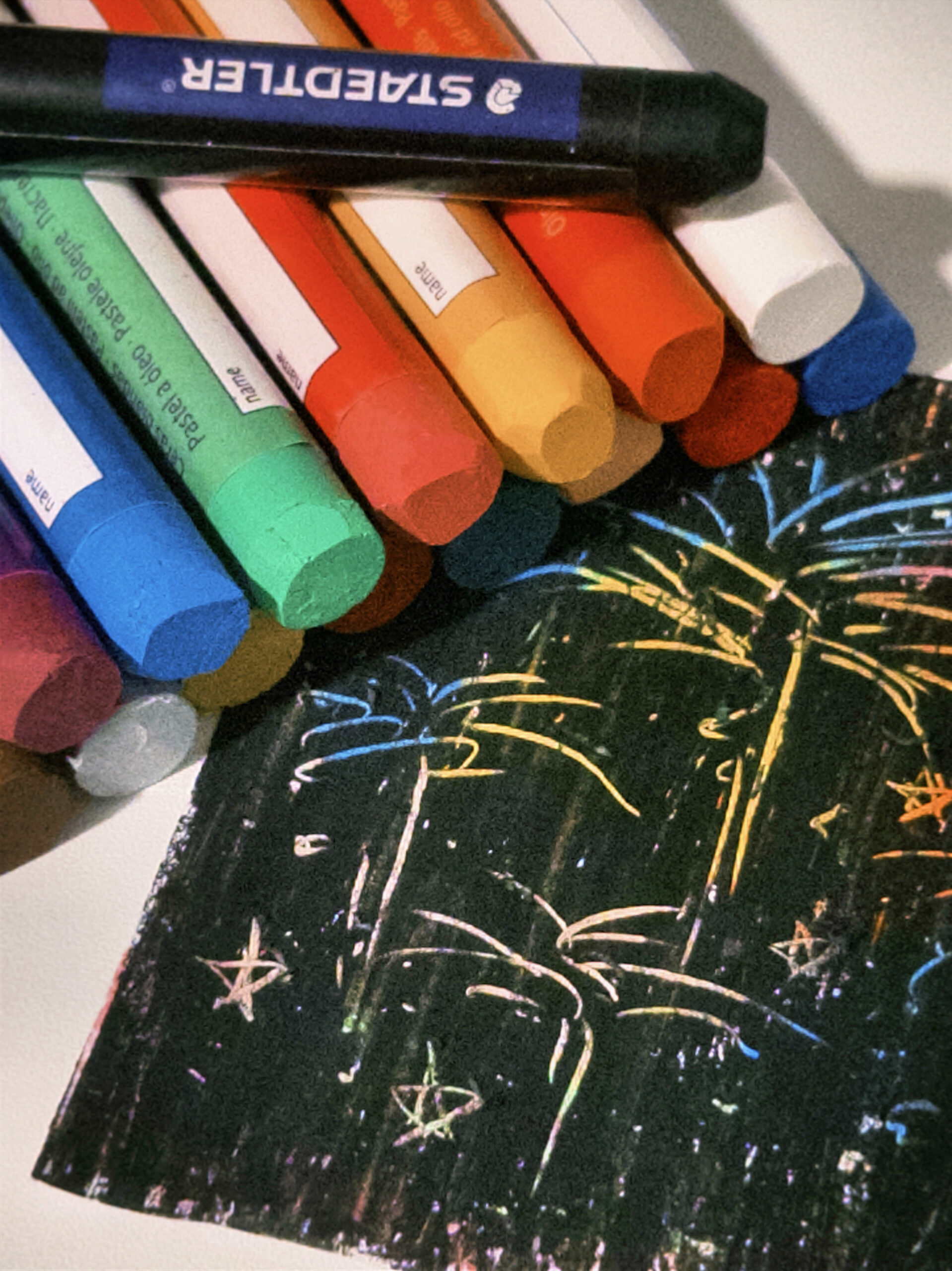 Cover of the toolkit including colored oil pastels, finished artwork and toolkit title. This is the cover of the toolkit. In the upper left corner of the image are two layers of colored oil pastels, with a black oil pastel placed horizontally on top. Bottom right is the result of this toolkit using oil pastels. This is a colorful firework bursting on a black background, with stars and various short lines surrounding it.