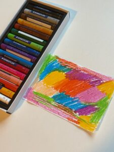 This is a piece of paper covered with oil pastels in various colors. To the left of the paper is a box of colored oil pastels.