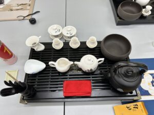 A black tea tray for holding all the items and draining excess waterA black kettle for heating water A white teapot for brewing tea A fair cup (gongdao cup) for pouring and equalizing the tea Several small tasting cups and fragrance cups A tea strainer and its holder A set of tea tools (tea scoop, tea tongs, etc.) in a black holder A brown rinse bowl for warming and cleaning cups A red tea towel for wiping the utensils A white pitcher and additional small bowls A packet of tea leaves on the side