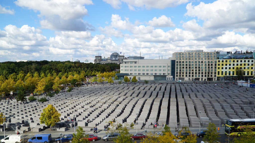 A large field of concrete stelae arranged in a grid in central Berlin, seen from above, surrounded by trees and buildings.