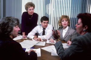 A group of office staff sitting around a table in a meeting, discussing documents — used to illustrate dimension 3 scenario.