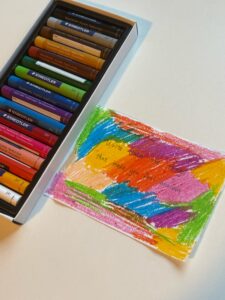 This is a piece of paper covered with various colors, and the colored oil pastel blocks cover the words below.To the left of the paper is a box of colored oil pastels.