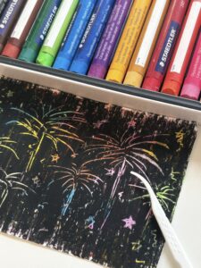 The paper covered with black oil pastels was scratched with fireworks, stars, and various short lines by toothpicks. On top of the paper is a box of colored oil pastels.