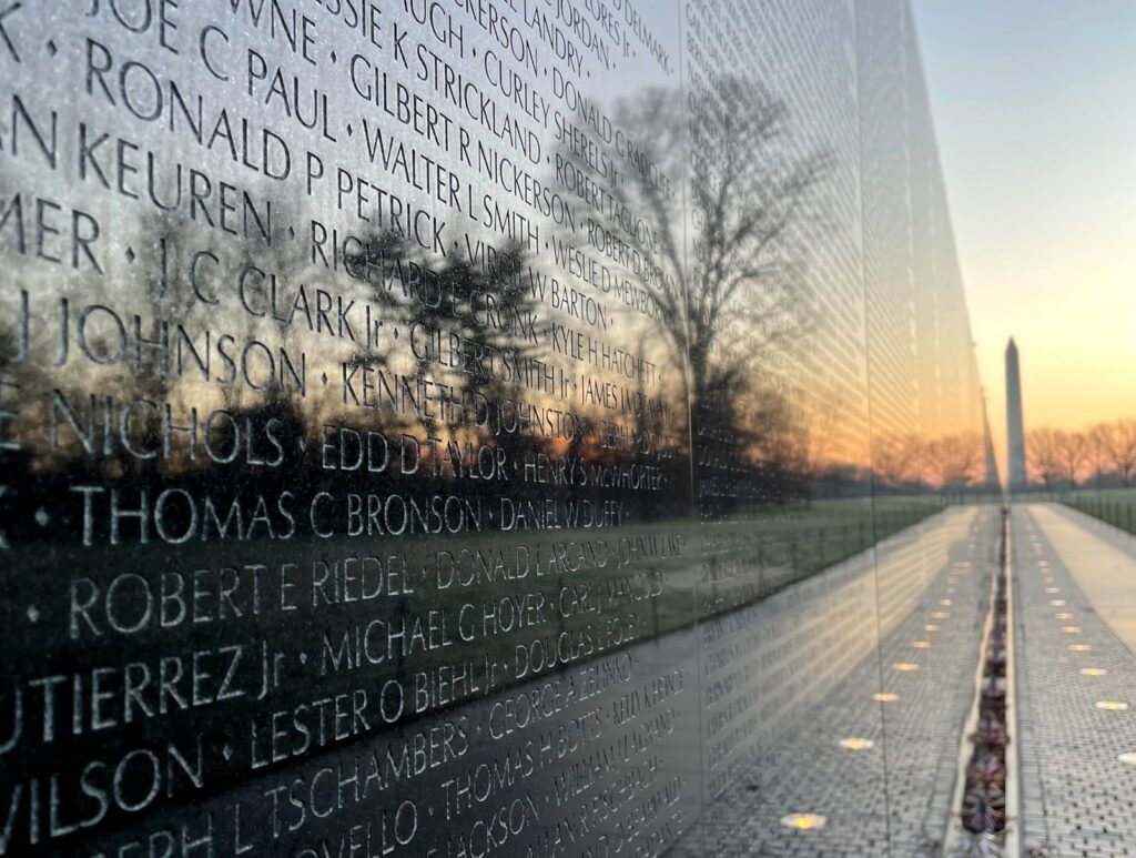 A close-up view of the Vietnam Veterans Memorial wall in Washington D.C., showing engraved names reflected in polished black granite at sunset. The Washington Monument is visible in the distance.