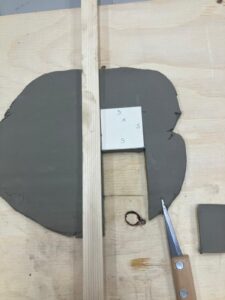 a flat piece of clay that has been cut into in a straight line using a potter's knife (also . pictured) There is a square template visible which is what will be used to guide the cutting of the clay.