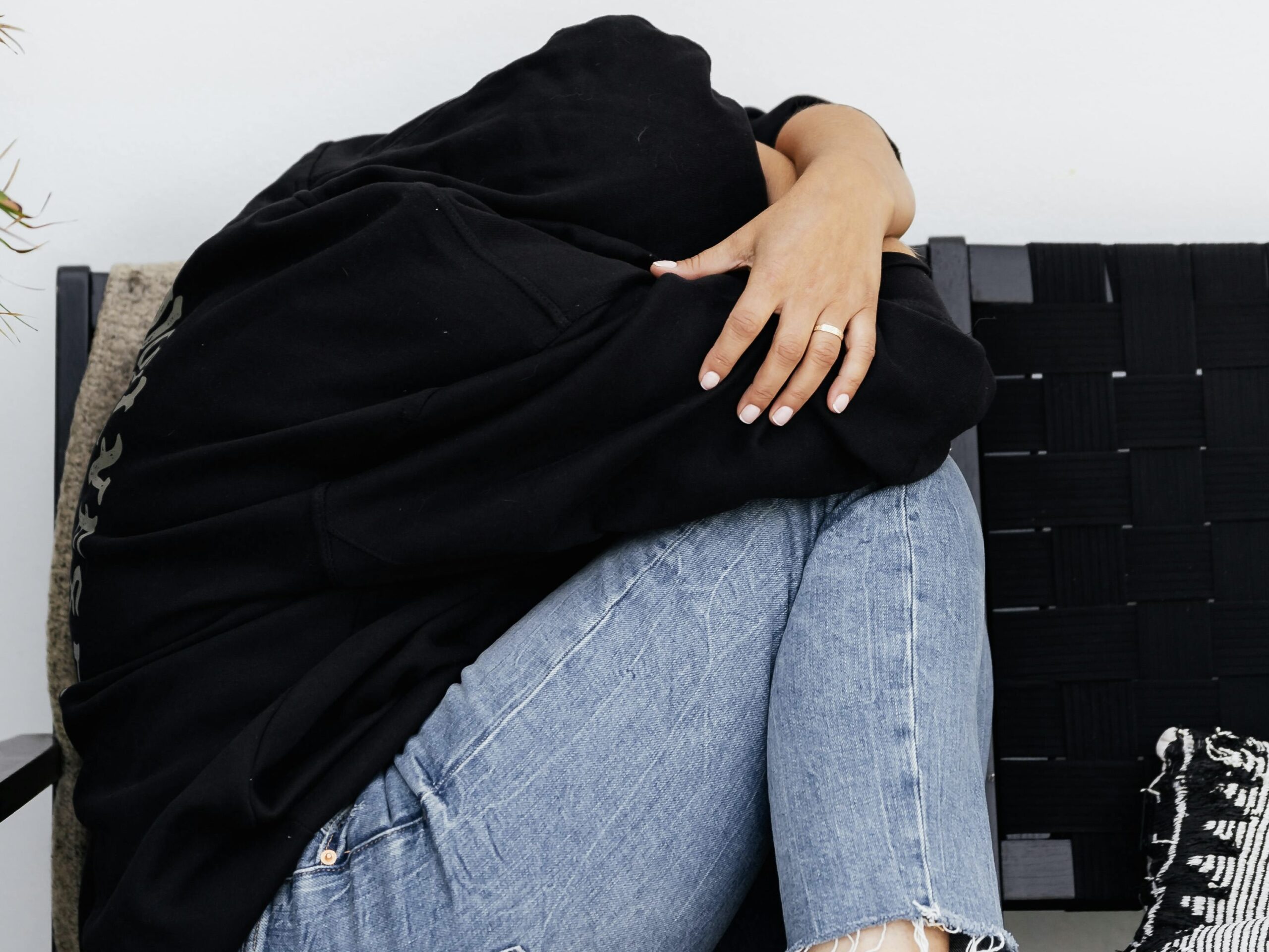 A person sits curled up on a chair, hugging their knees while wearing a black hoodie and light jeans.