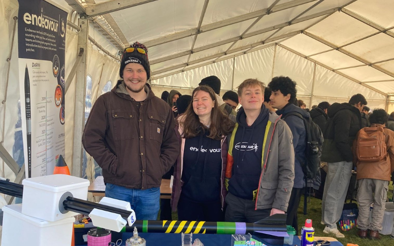 Three students at information desk with students and pupils in crowd behind. All instead a large white tent.