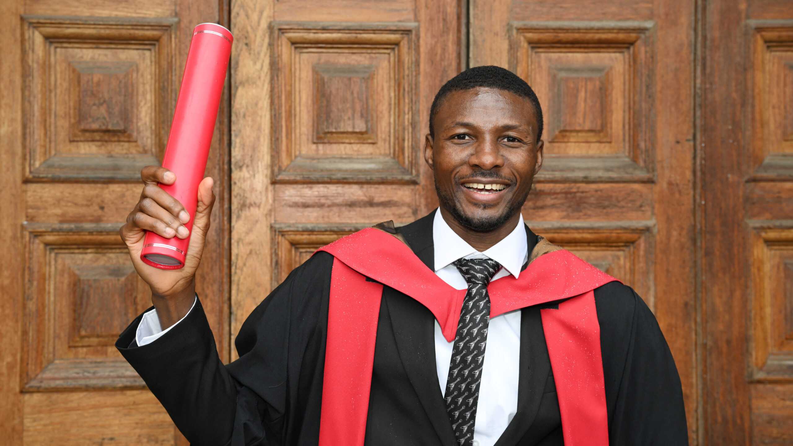 Graduate proudly holding up his red graduation scroll outside ornate wooden doors.