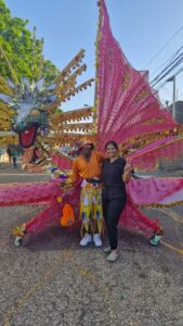 Arrianna Peters smiles and makes a peace sign beside a carnival performer in an elaborate orange and gold costume, with large decorative pink and gold carnival wings in the background.