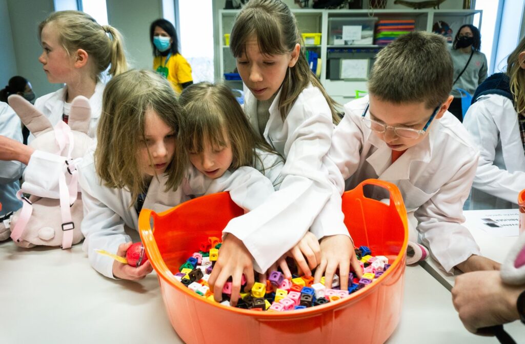 Children wearing lab coats group around a bucket containing plastic building blocks during the Science Festival 2022