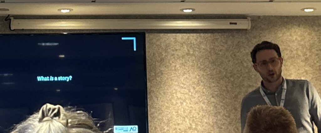 A picture of Stewart Utley, a white man with glasses wearing a grey button down, beside his powerpoint slide asking "What is a story?"