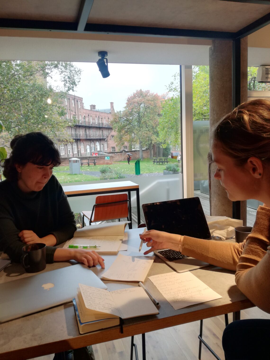 Two white women sit at a table talking and pointing at notes. Two laptops are visible suggesting a working environment. In the background through an open window you can see trees and a red brick building