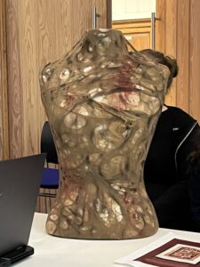 A torso-shaped sculpture sits on a table in a classroom setting, covered with layered textures and embedded circular forms, with wood-paneled doors and seated students in the background.