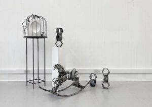 Installation view in a white studio: at left, a tall black metal stand supports a circular metal cage containing a white block with a balloon-shaped cavity. Centre foreground, a stainless-steel rocking-horse-like sculpture rests on curved runners. To the right, two small modular steel figures stand on the floor, and one similar figure sits on a white plinth.