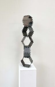 A tall, vertical sculpture on a white plinth. Dark metal polygonal frames interlock like modular joints, forming a totem-like figure against a plain wall.