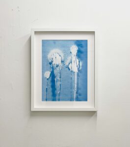 A framed blue artwork on a white wall. The image shows pale, vertical forms with dripping marks, like ghostly figures emerging from washes of pigment.