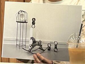 A person holds a printed photograph showing a metal sculpture installation in a studio space, with several small sculptural forms arranged on the floor and a tall stand at the left.