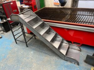 A metal staircase structure in a workshop environment. The steps are silver sheet metal with welded seams; red machinery and industrial surfaces surround it.