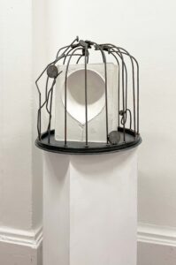 A round metal cage sits on a circular base atop a white plinth. Inside is a white block with a smooth, balloon-shaped cavity. Thin metal rods arch over the form.