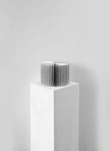 A small sculptural book sits on a tall white plinth against a pale wall. The pages are fanned into a circular or cylindrical form, creating a dense, textured surface.