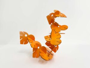 A small orange modular sculpture on a white surface. Flat, slotted petal-like pieces form a curved line, arching upward at one end and bending down at the other.