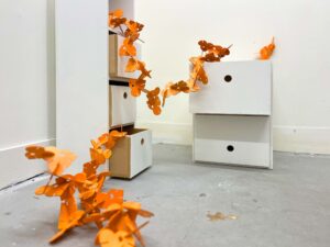 An installation view in a white room: orange modular forms snake from the floor up into open drawers and across to a small cabinet. The pieces appear to “crawl” through the furniture.