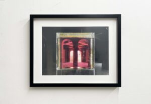 A black-framed artwork on a white wall. Inside the frame is an image dominated by deep reds and pinks, showing an architectural interior with arched shapes and light reflections.
